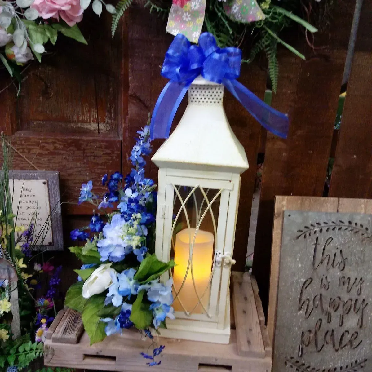 White lantern with a glowing candle, blue flowers, and a blue bow on a wooden crate.