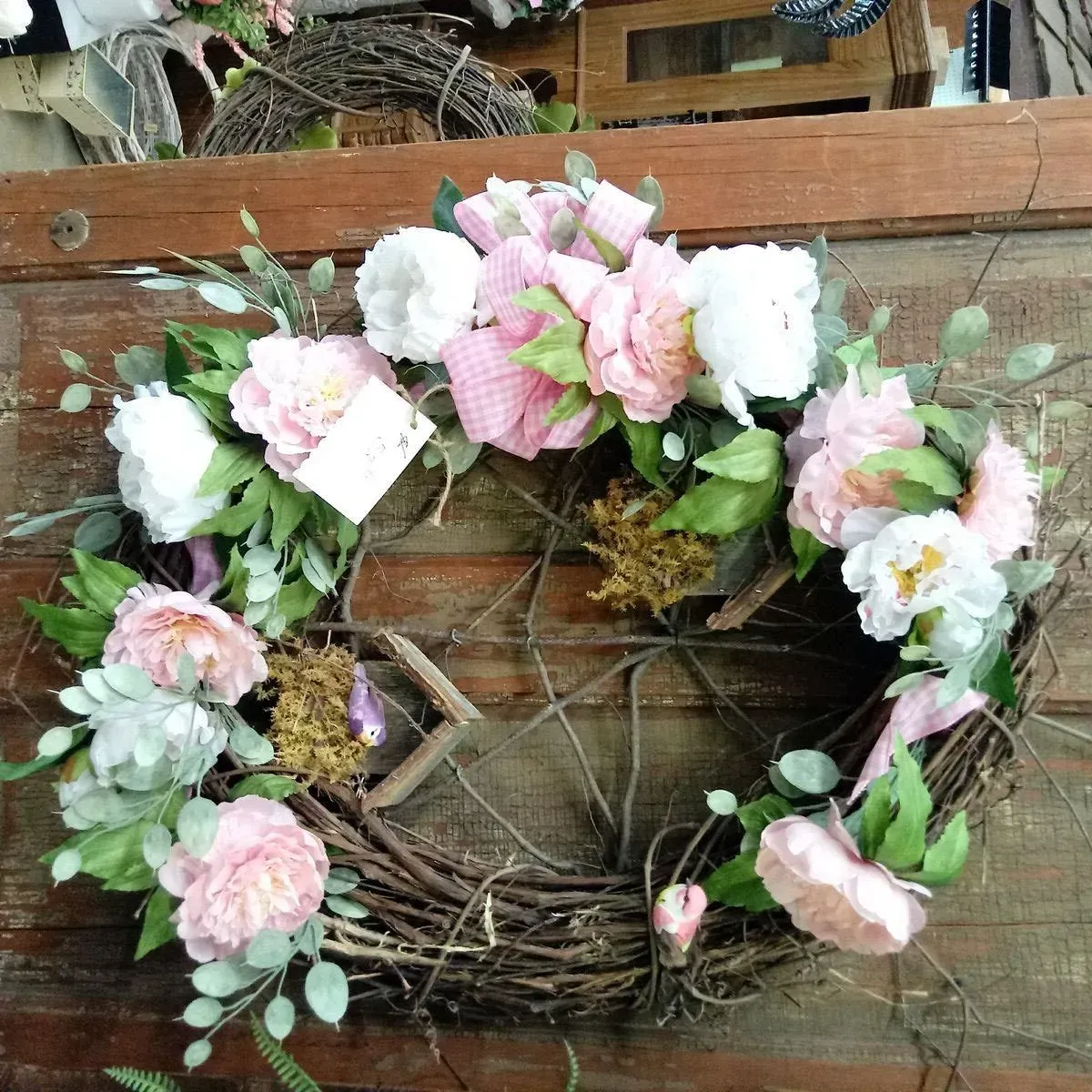 Wreath made of twigs, adorned with pink and white flowers, green leaves, and a pink gingham ribbon.