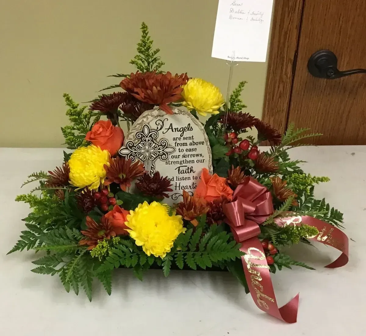 Floral arrangement with a stone plaque. Yellow, orange, and maroon flowers with a burgundy ribbon, set against a wooden background.