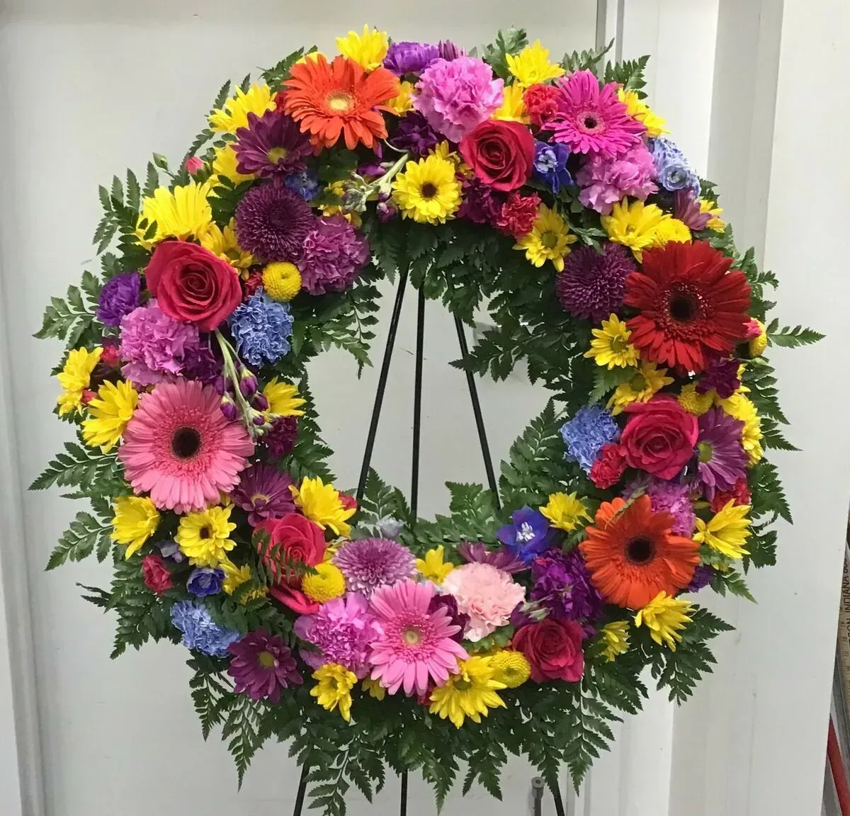 Colorful floral wreath on a black stand, featuring various flowers in shades of pink, yellow, red, purple, and blue.
