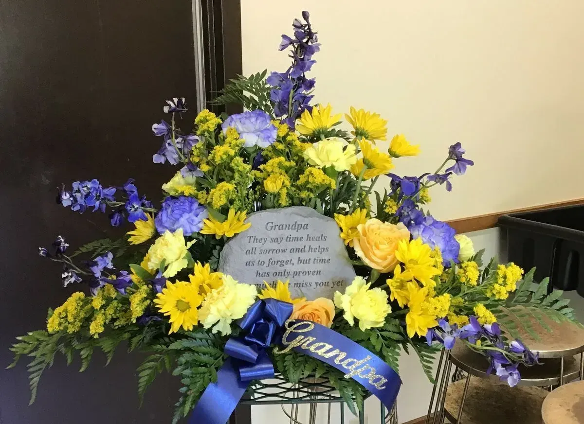 Floral arrangement with blue and yellow flowers, a memorial stone, and a blue ribbon that says Grandpa.
