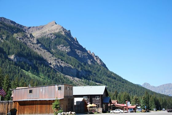Wooden buildings sit in front of a mountain covered in trees under a clear blue sky.