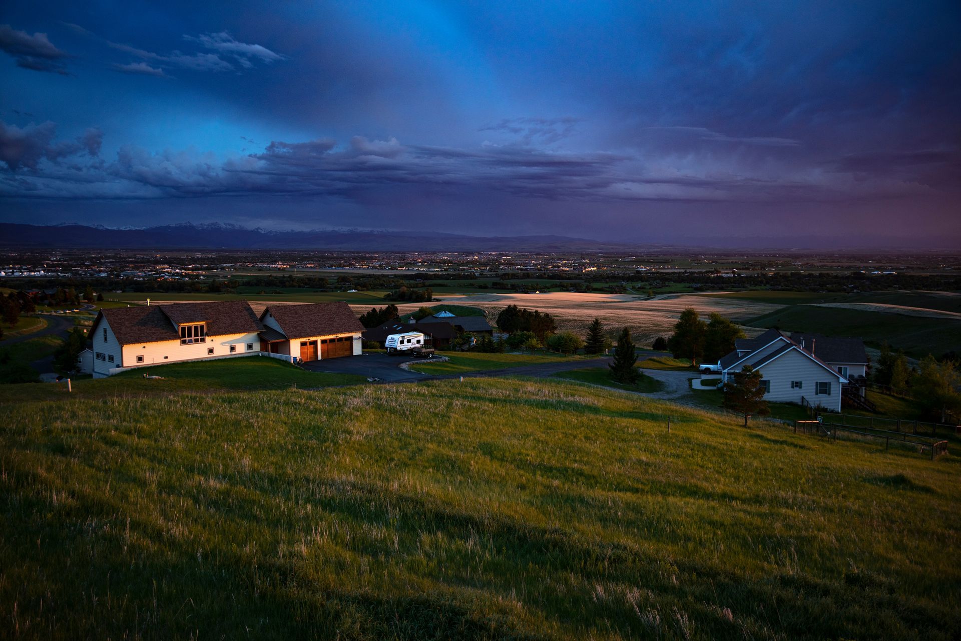 Houses on a grassy hill at dusk, city lights in the distance under a blue and purple sky.