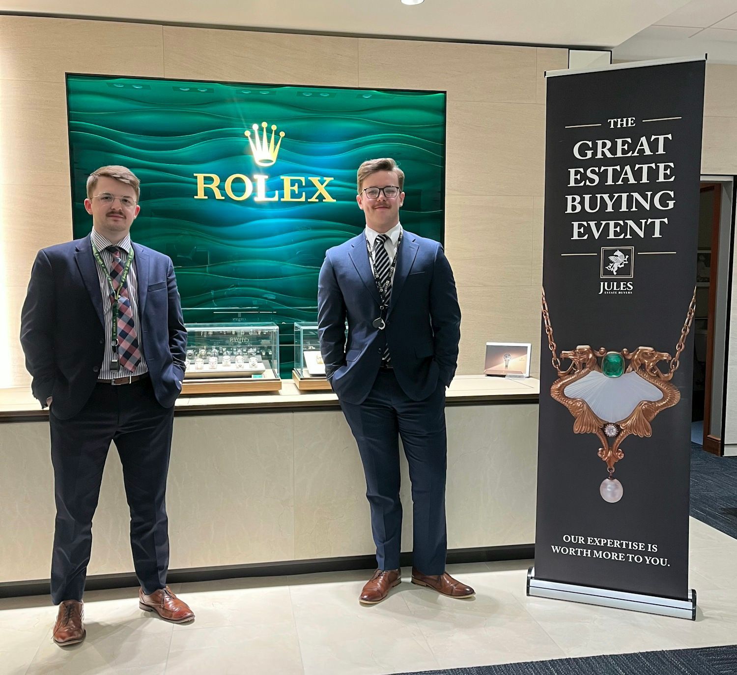 Two men in suits stand by a Rolex display and a banner for 