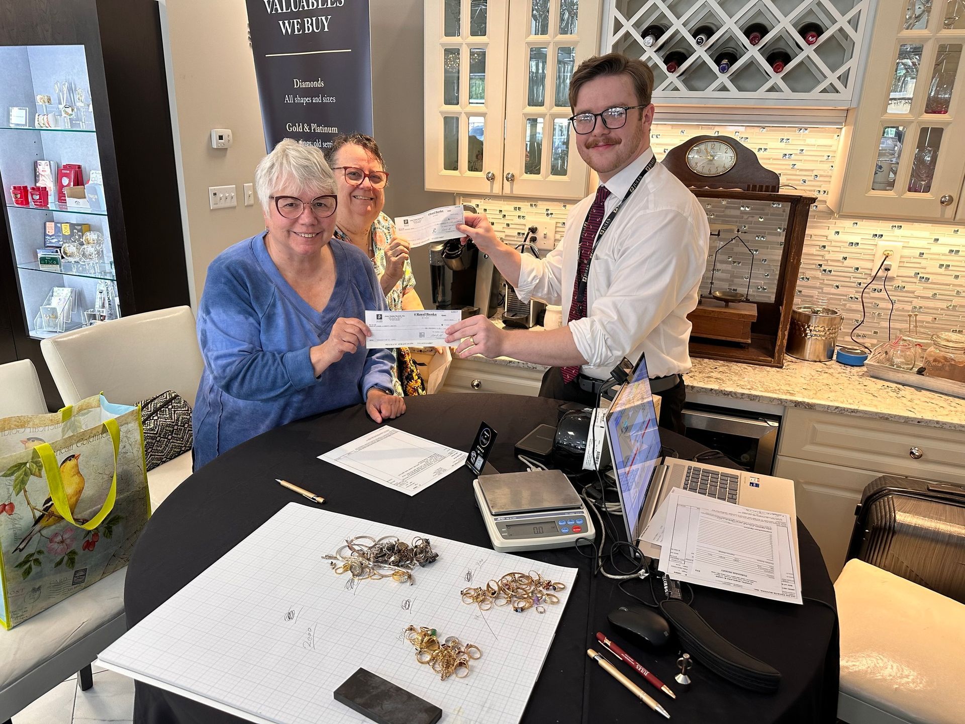 A man in a tie hands a check to a woman; another woman smiles nearby. The scene takes place indoors at a jewelry store with gold items on a table.