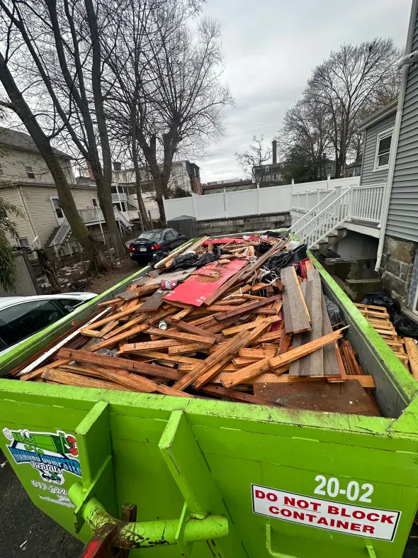 A green dumpster filled with wood is parked in front of a house.