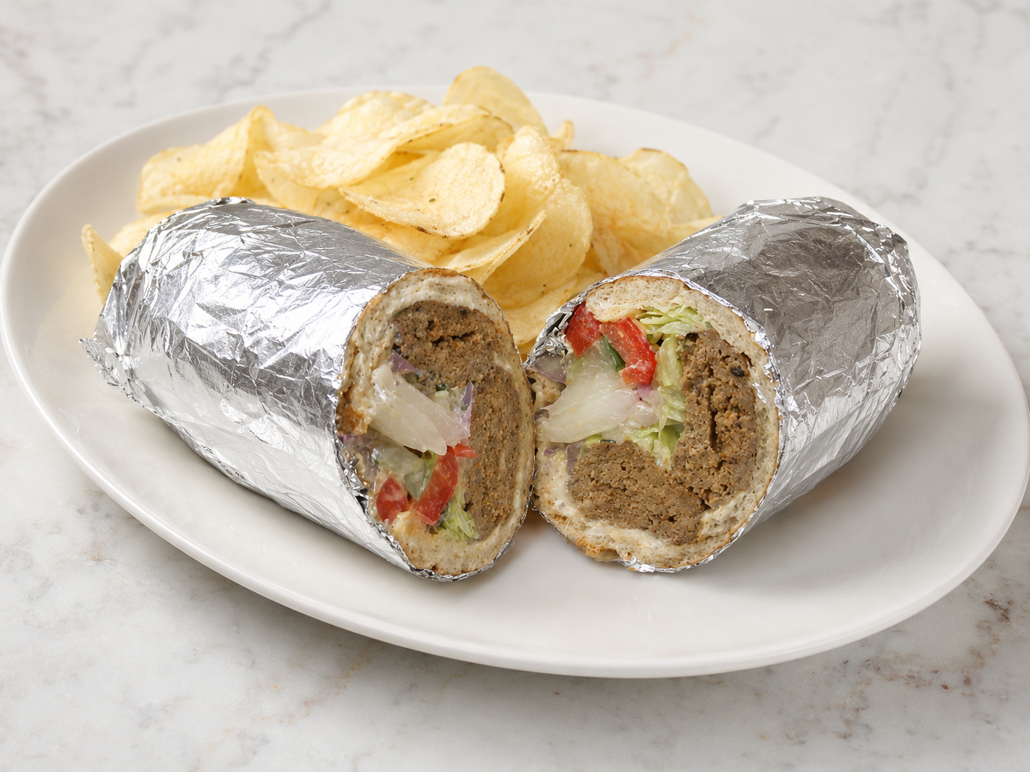 Foil-wrapped burrito cut in half on a plate with potato chips in the background
