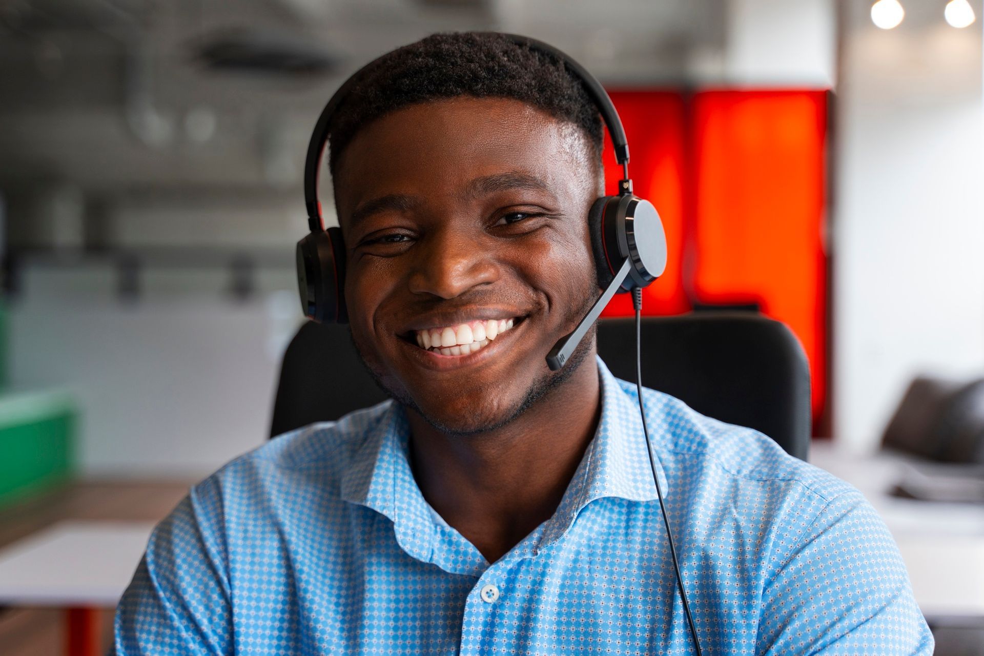 Smiling Black man wearing a headset, blue shirt, seated in an office setting.