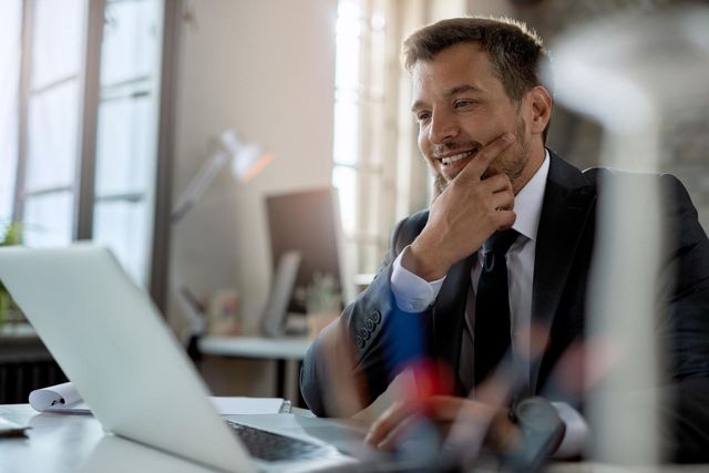 Businessman smiling at a laptop in a bright office, hand on chin