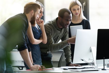 Four diverse colleagues collaborating, looking at a computer screen in a modern office setting.