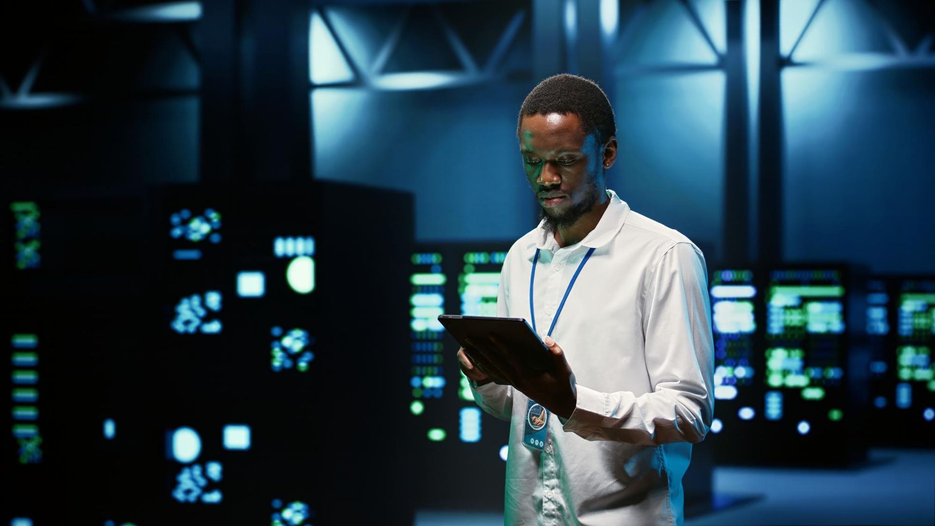 Man in white shirt using a tablet in a dimly lit data center with glowing monitors