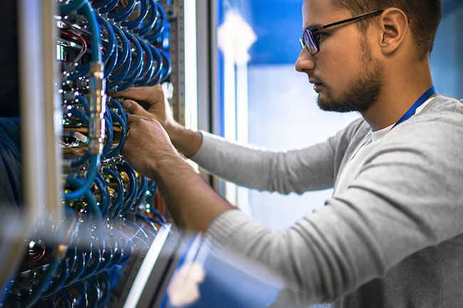A man in glasses connects cables in a server room, wearing a gray sweater.