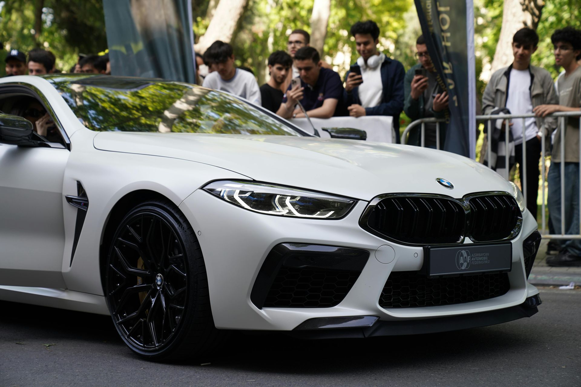 White sports car parked outdoors at a car show, with people standing behind barriers.