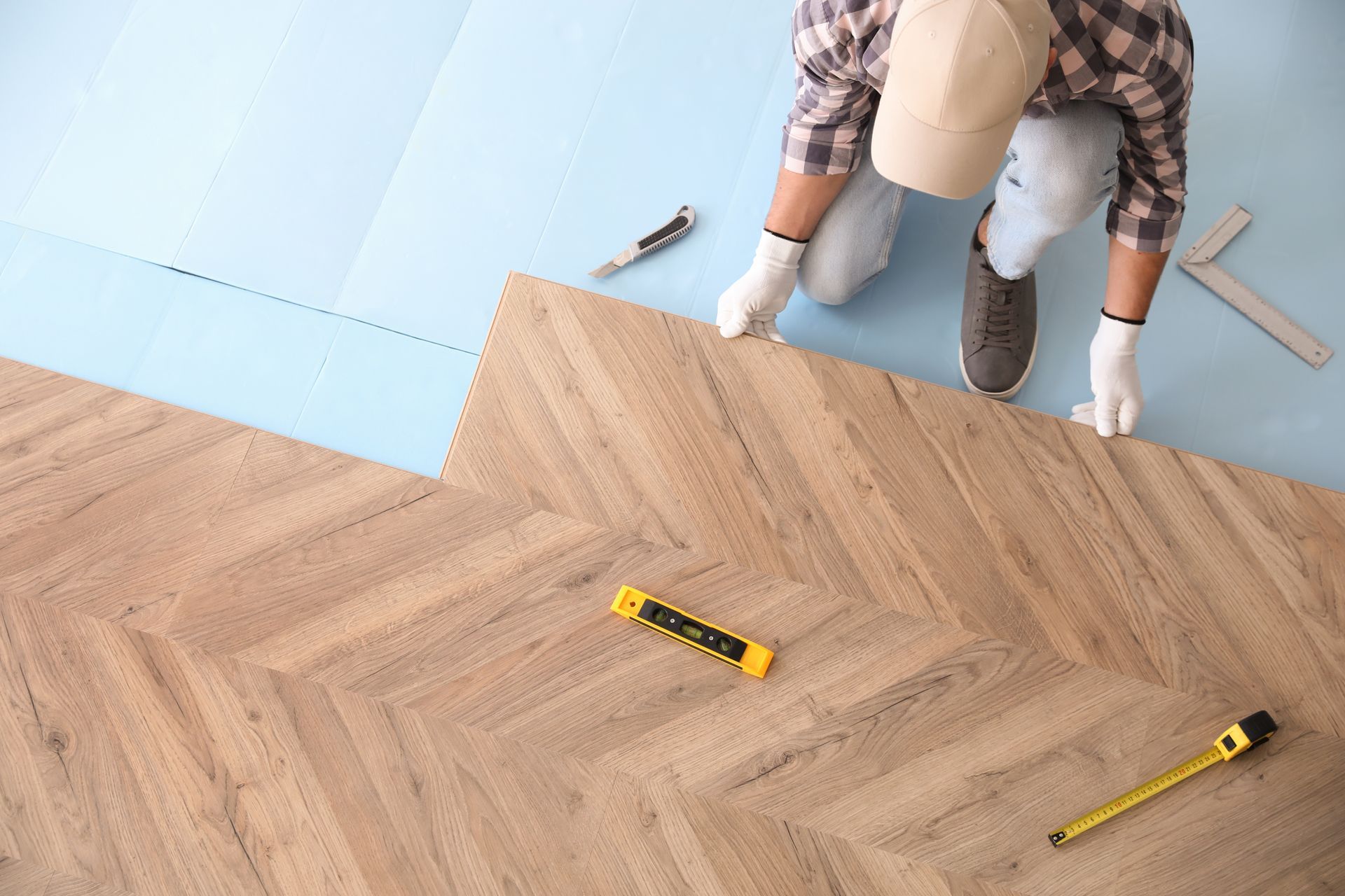 A worker installs herringbone-patterned wood flooring over a light blue underlayment, using tools on the floor.