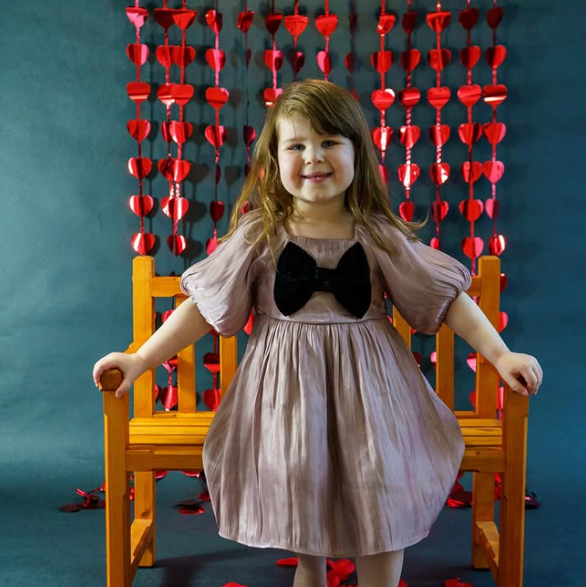 A little girl in a pink dress with a black bow is standing on a chair