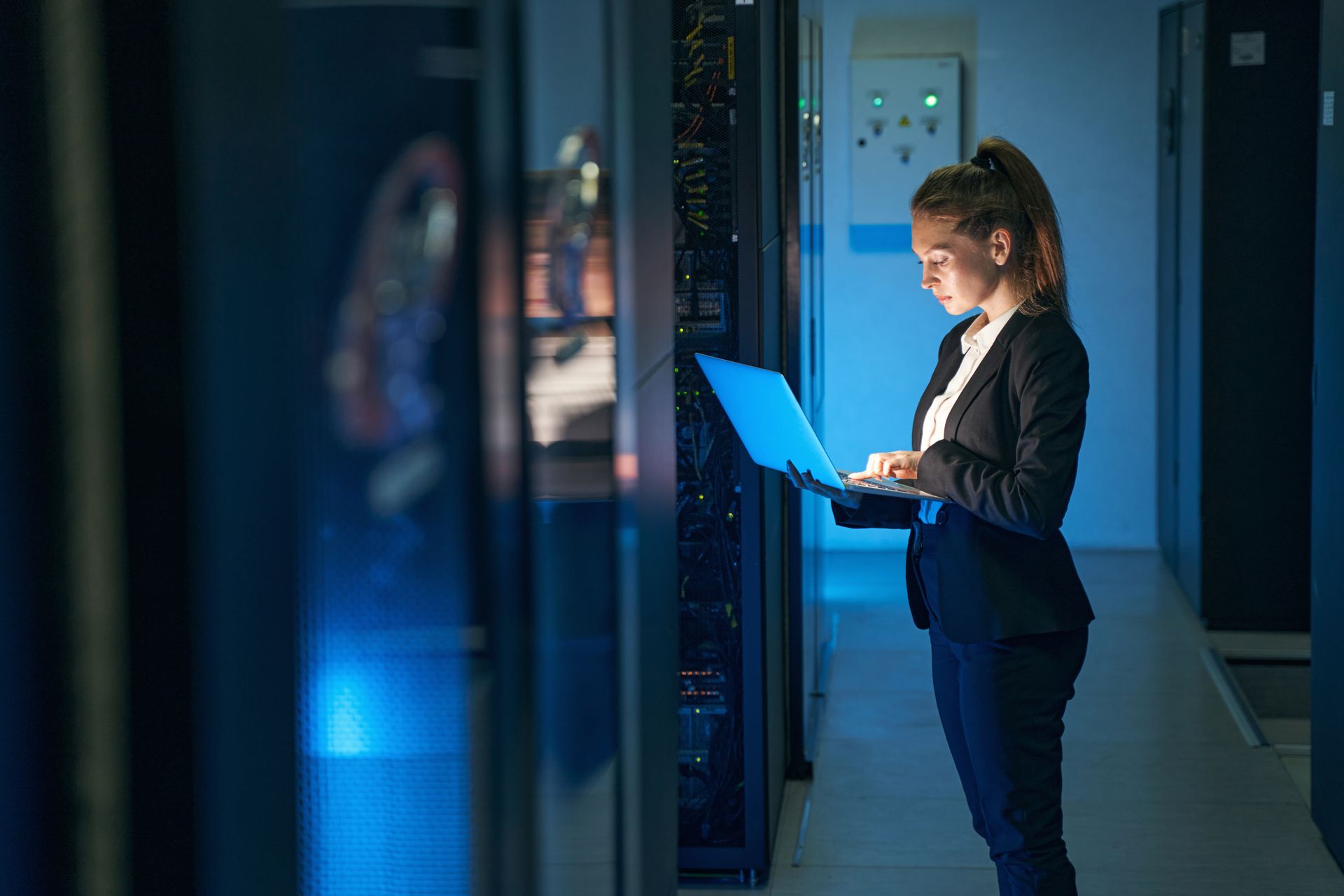 A woman is standing in a server room using a laptop computer.