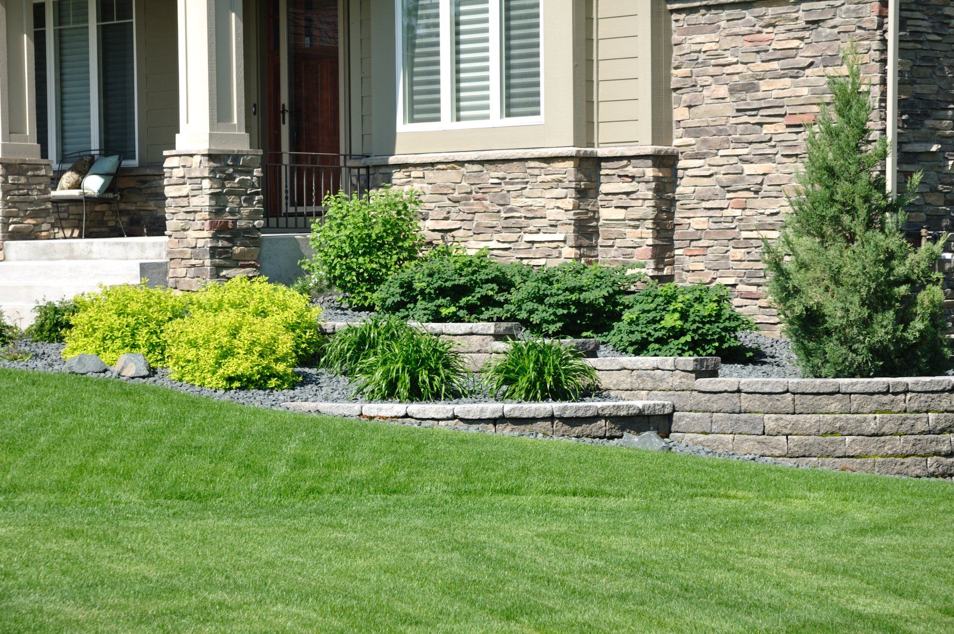 Landscaped front yard with stone retaining wall, shrubs, flowers, and green lawn beside a house