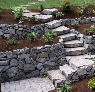Tiered stone garden wall with steps, gray pavers, and small green plants on a sloped landscape