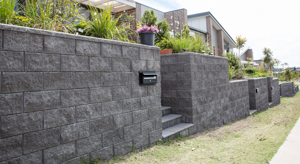 Dark stone retaining wall with steps and mailbox along a grassy roadside beside homes