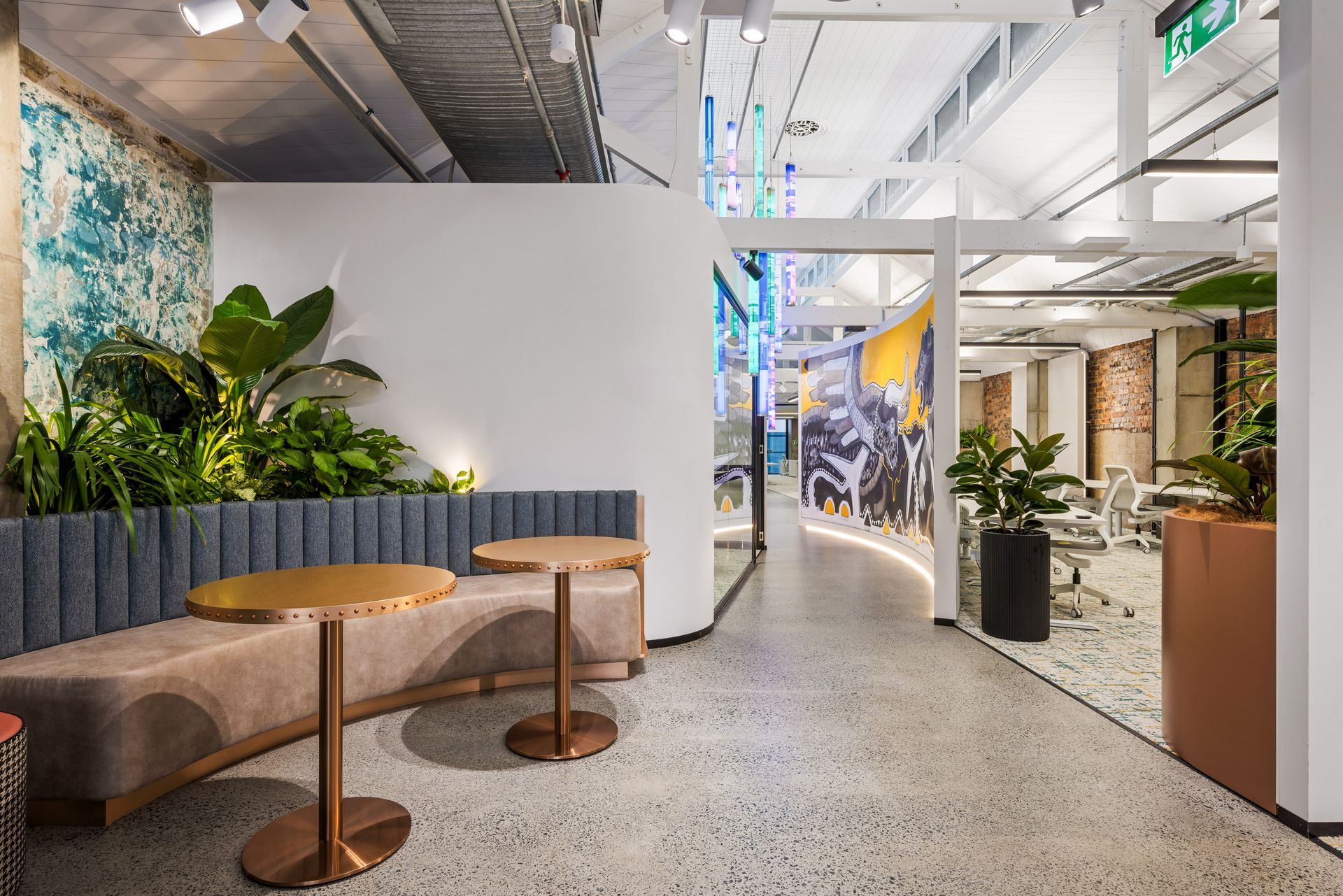 Modern office lounge with round tables, cushioned seating, and plants along a bright corridor
