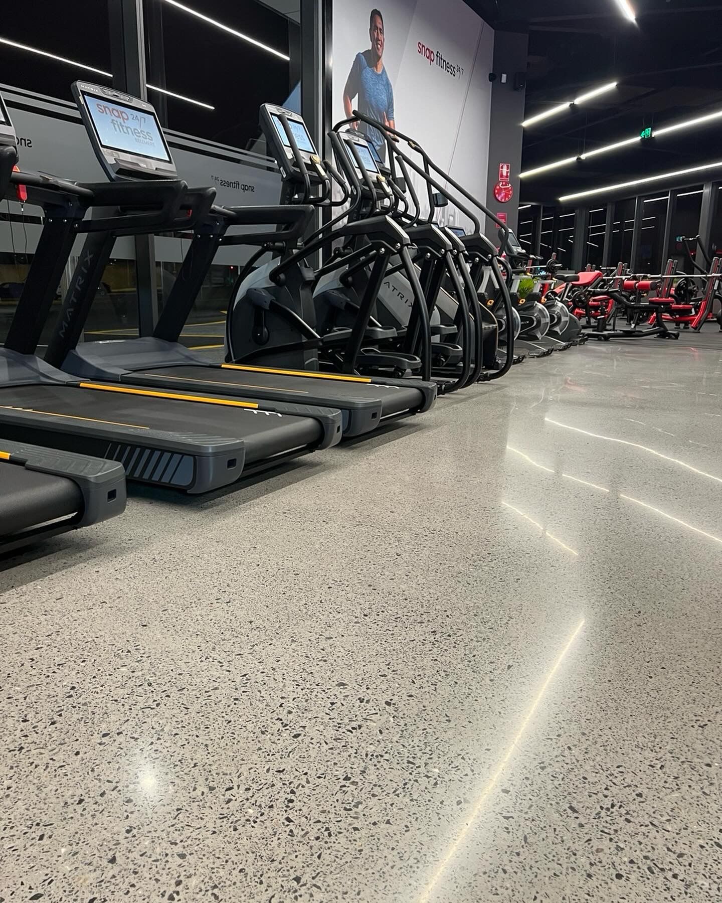 Gym with treadmills and bikes under bright lights, viewed from a low angle across a speckled floor