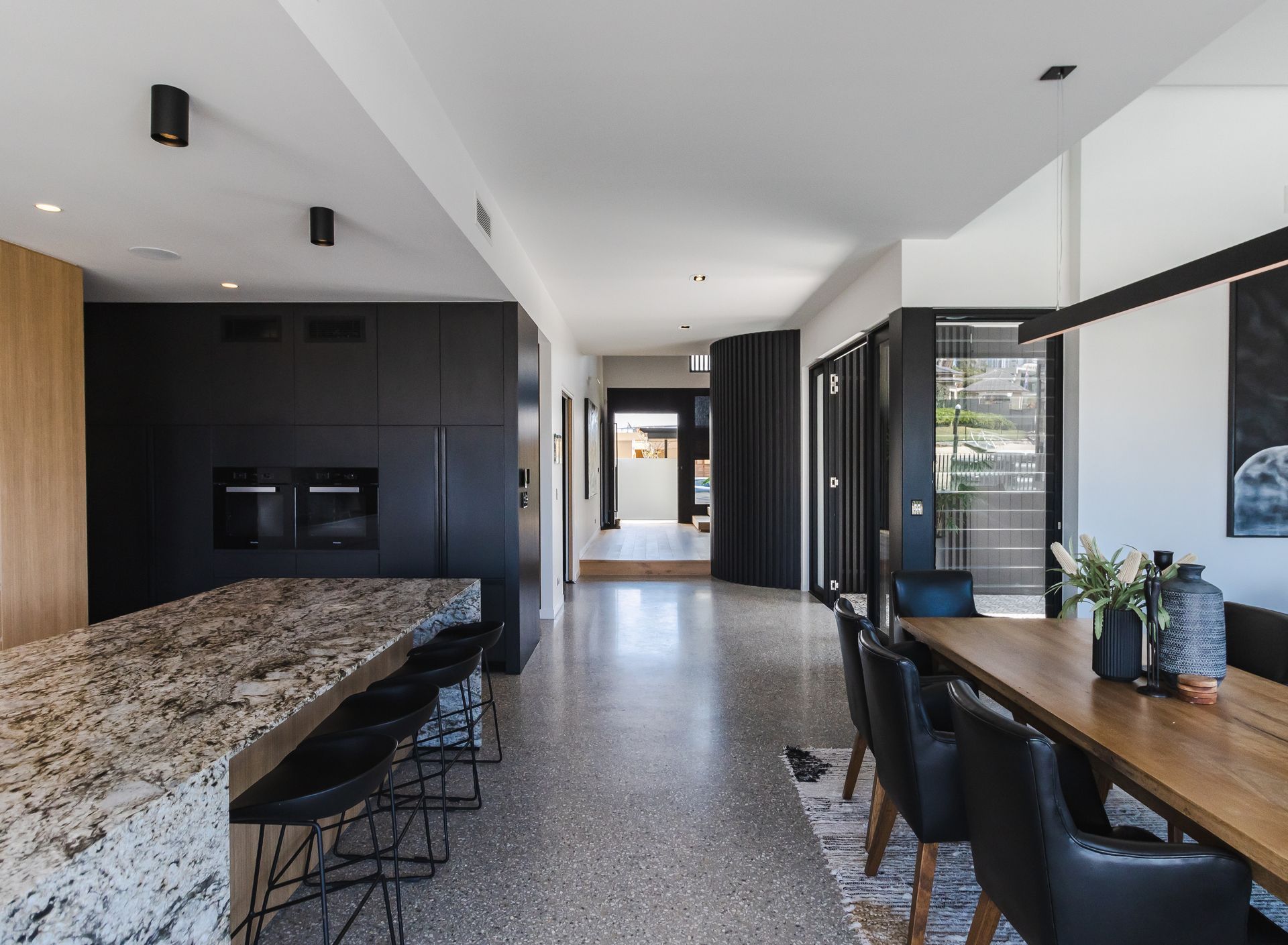 Modern open-plan kitchen and dining area with black accents, granite island, and terrazzo floor leading to a hallway