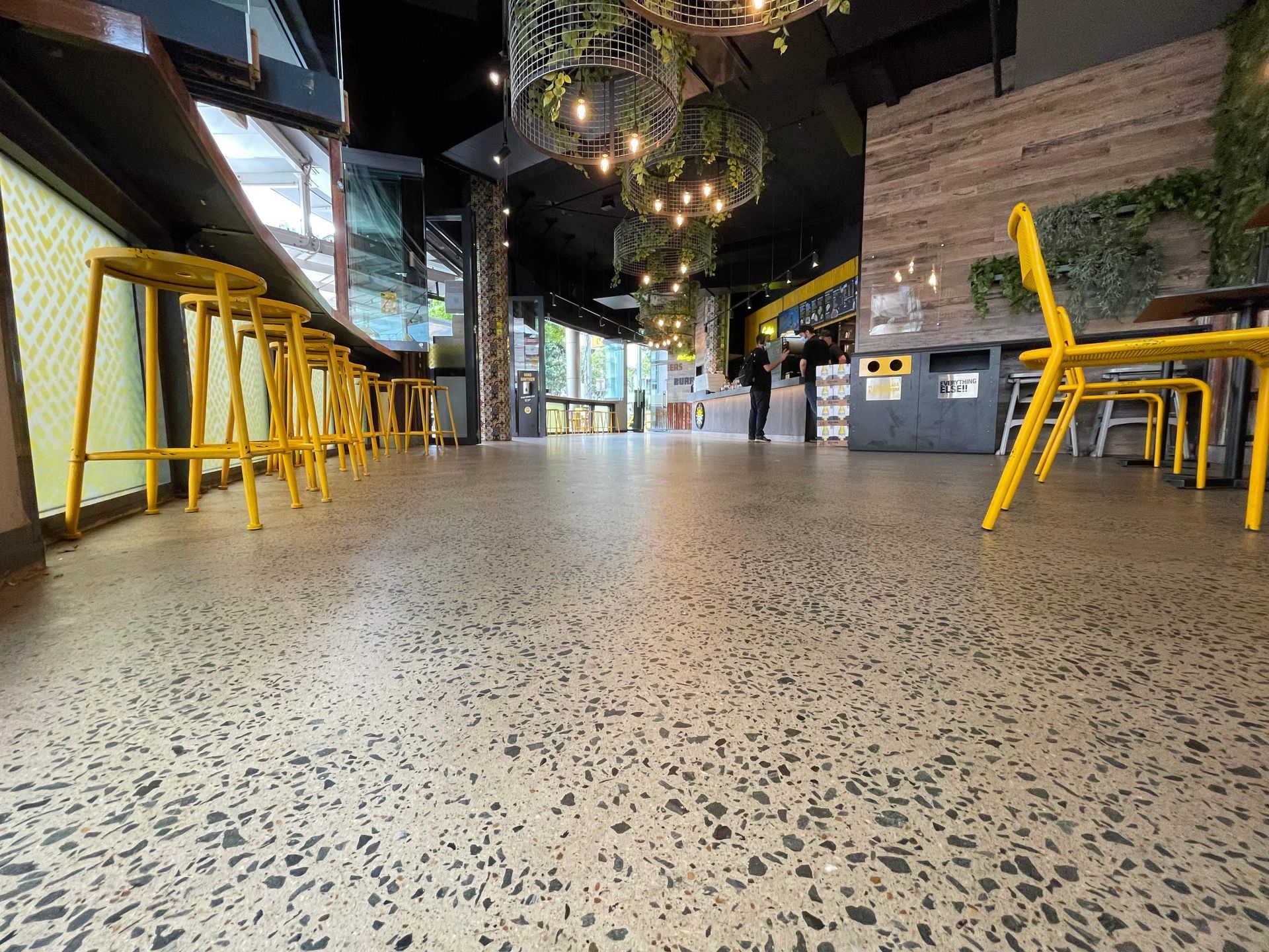Long café interior with yellow bar stools, speckled floor, and hanging lights.