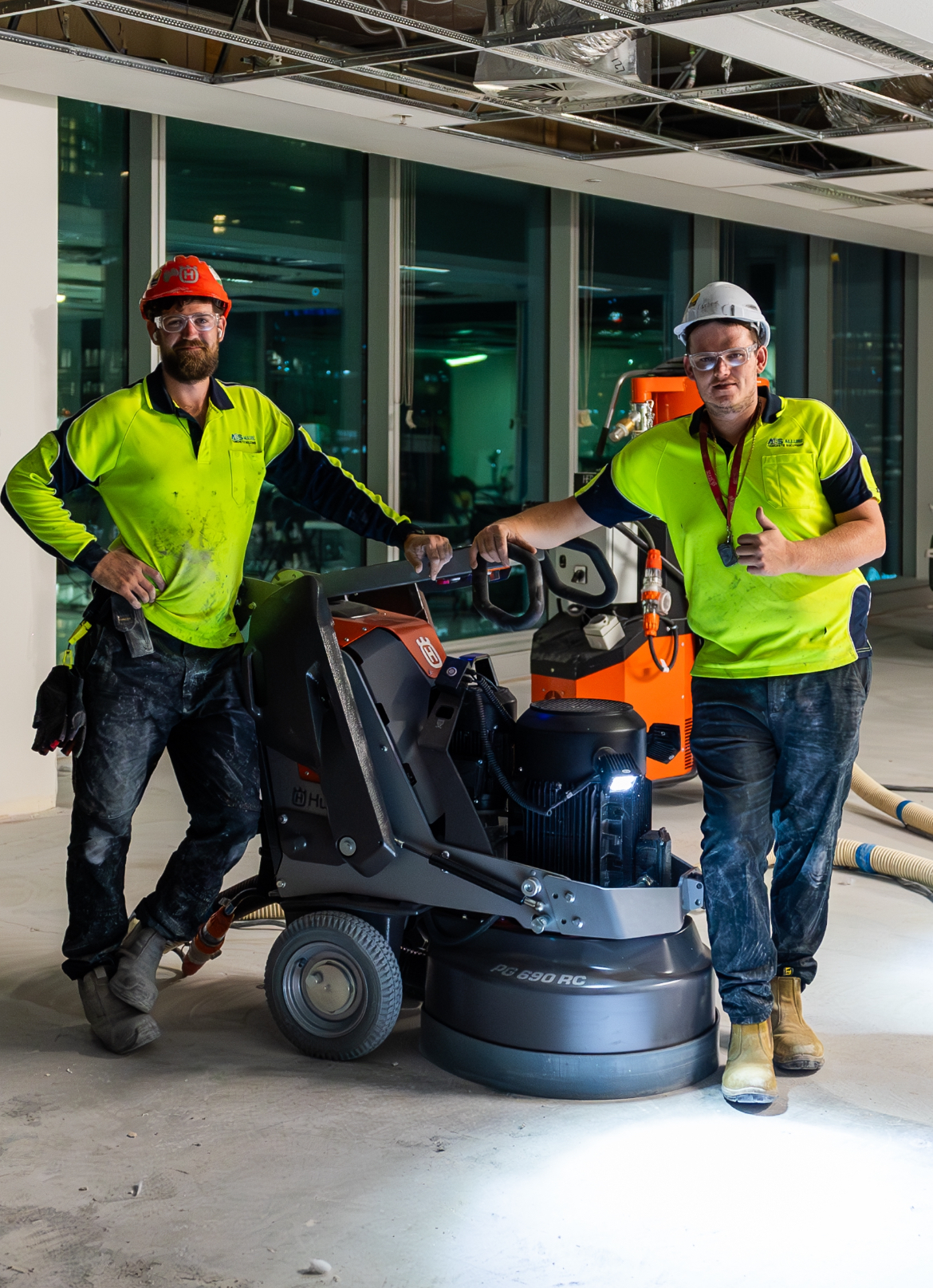 Two workers in neon safety shirts posing with a floor scrubber in an indoor construction site.