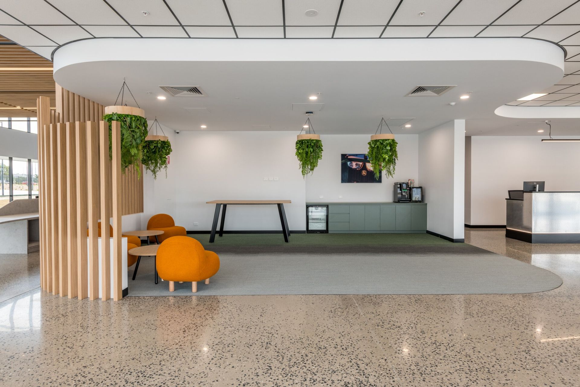 Modern office lounge with orange chairs, green plants, and a long gray bench under white curved ceiling panels