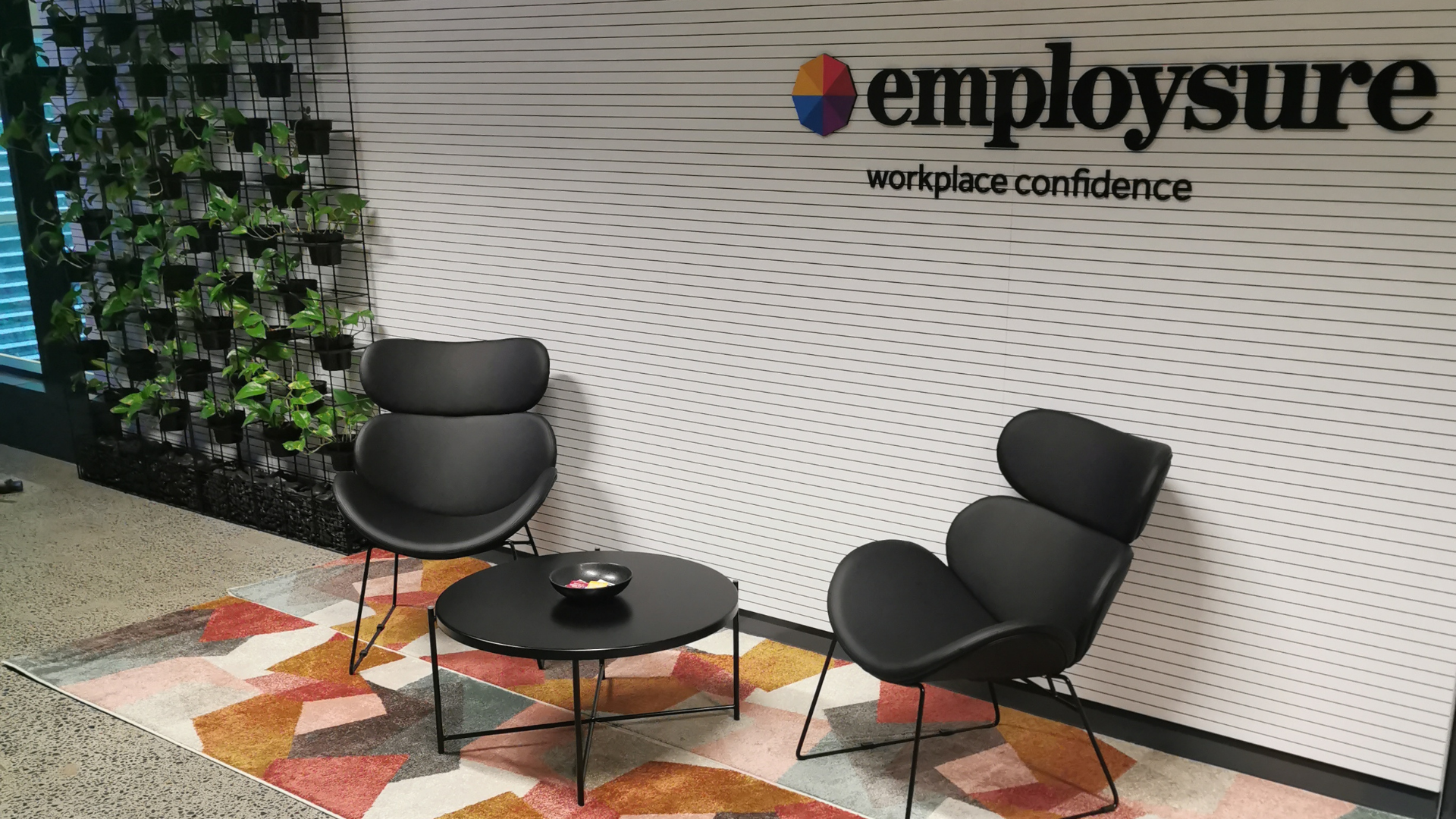 Modern office lounge with two black chairs, small round table, plant wall, and Employsure branding on the wall Modern office lounge with two black chairs, small round table, plant wall, and Employsure branding on the wall