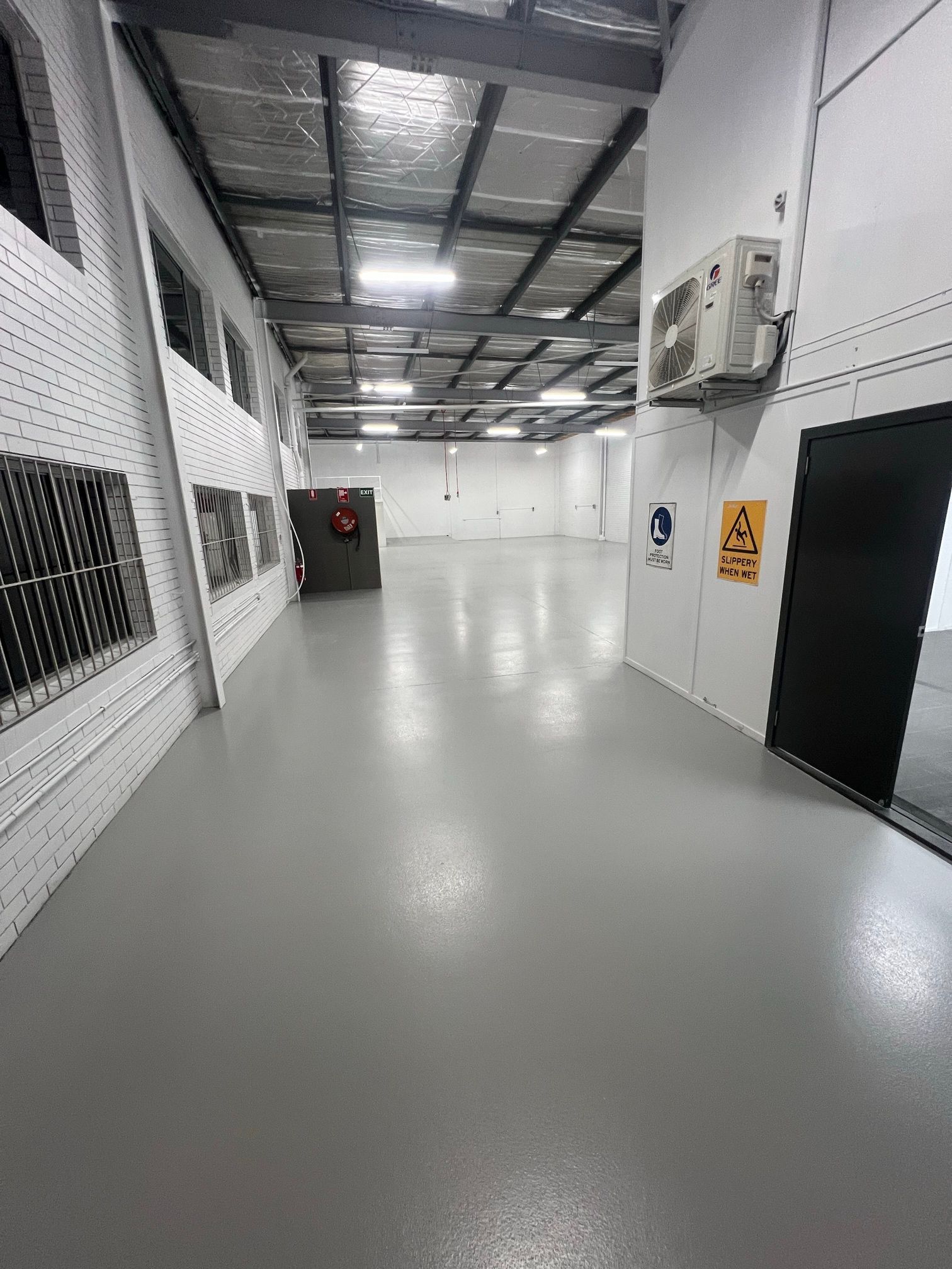 Empty industrial hallway with white walls, gray floor, and ceiling lights, showing a black door and wall signs.