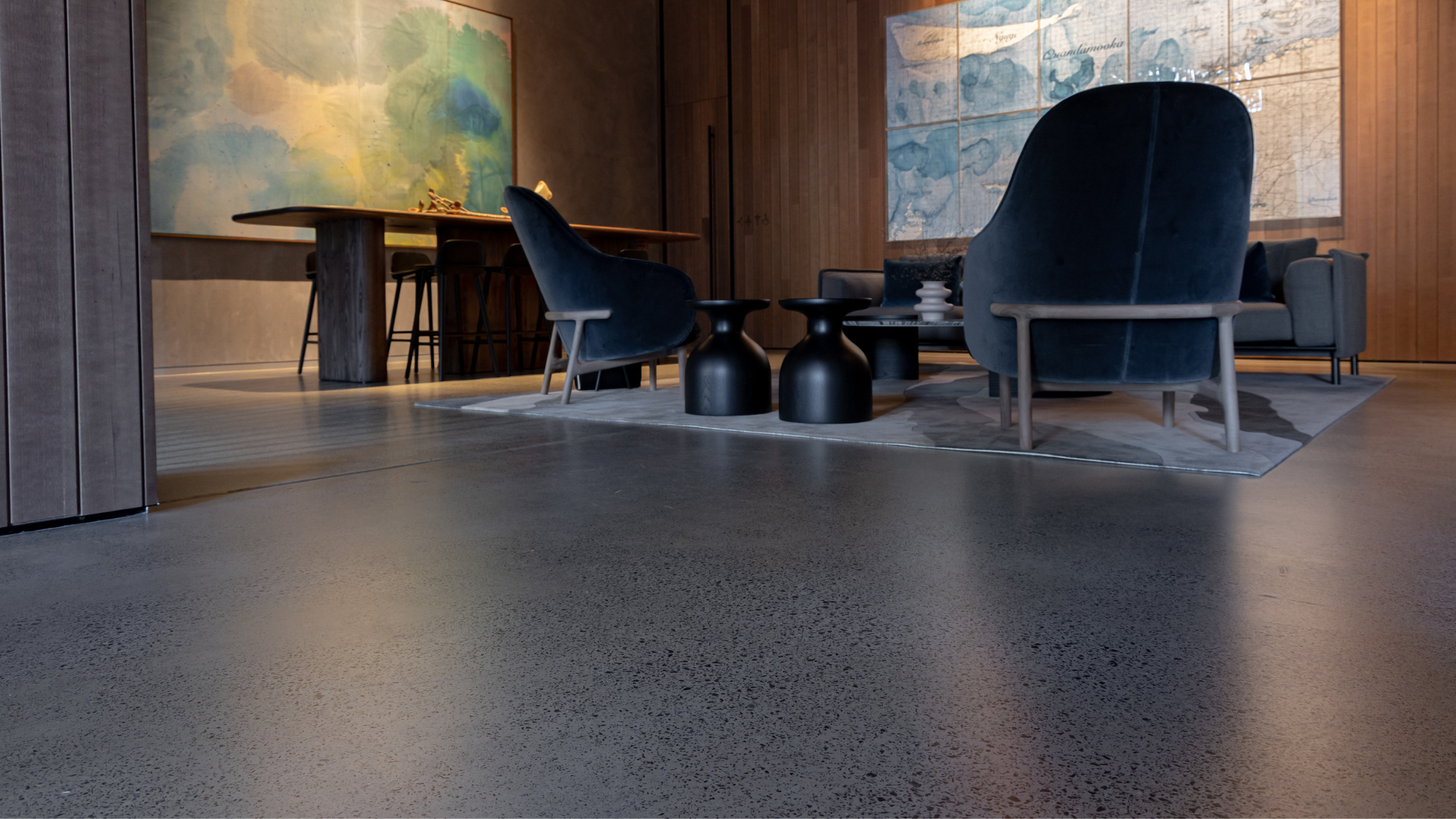 Modern lounge with navy chairs, low black tables, warm wood walls, and abstract art behind a polished concrete floor.