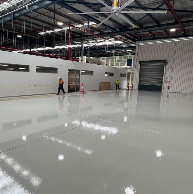 Workers in a bright industrial warehouse with a glossy white floor and overhead lights