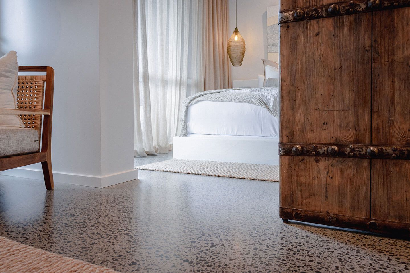 Minimal bedroom with white bed, sheer curtains, hanging pendant light, and rustic wood door on terrazzo floor