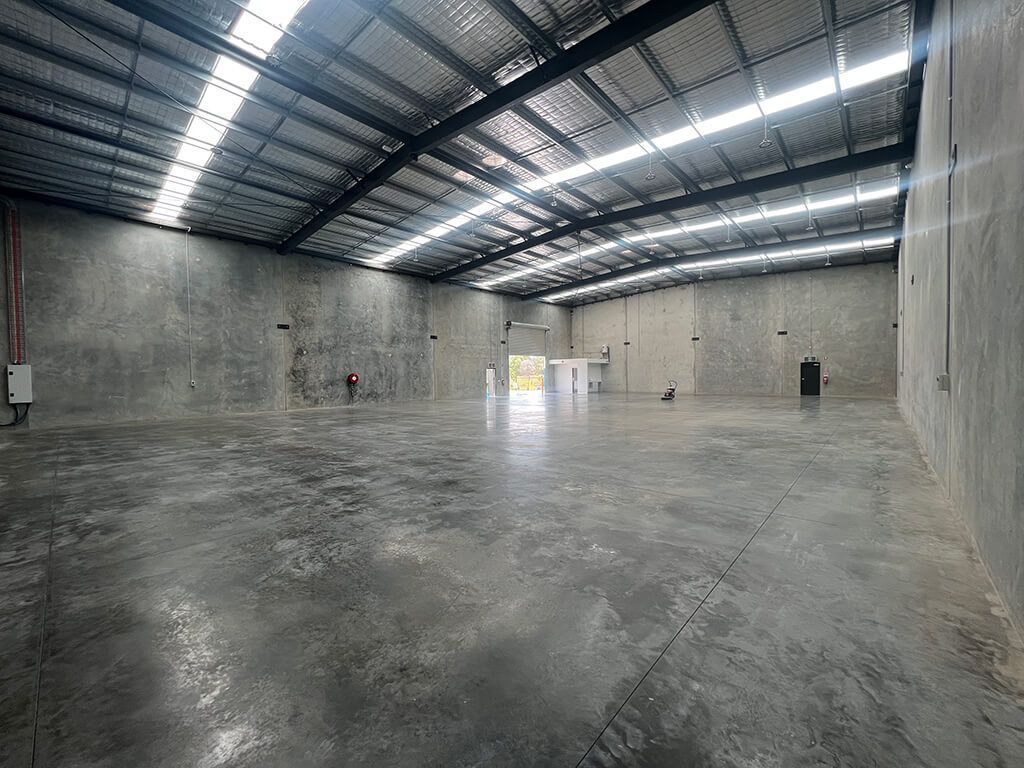 Empty concrete warehouse with high metal roof and glossy floor, lit by daylight through skylights