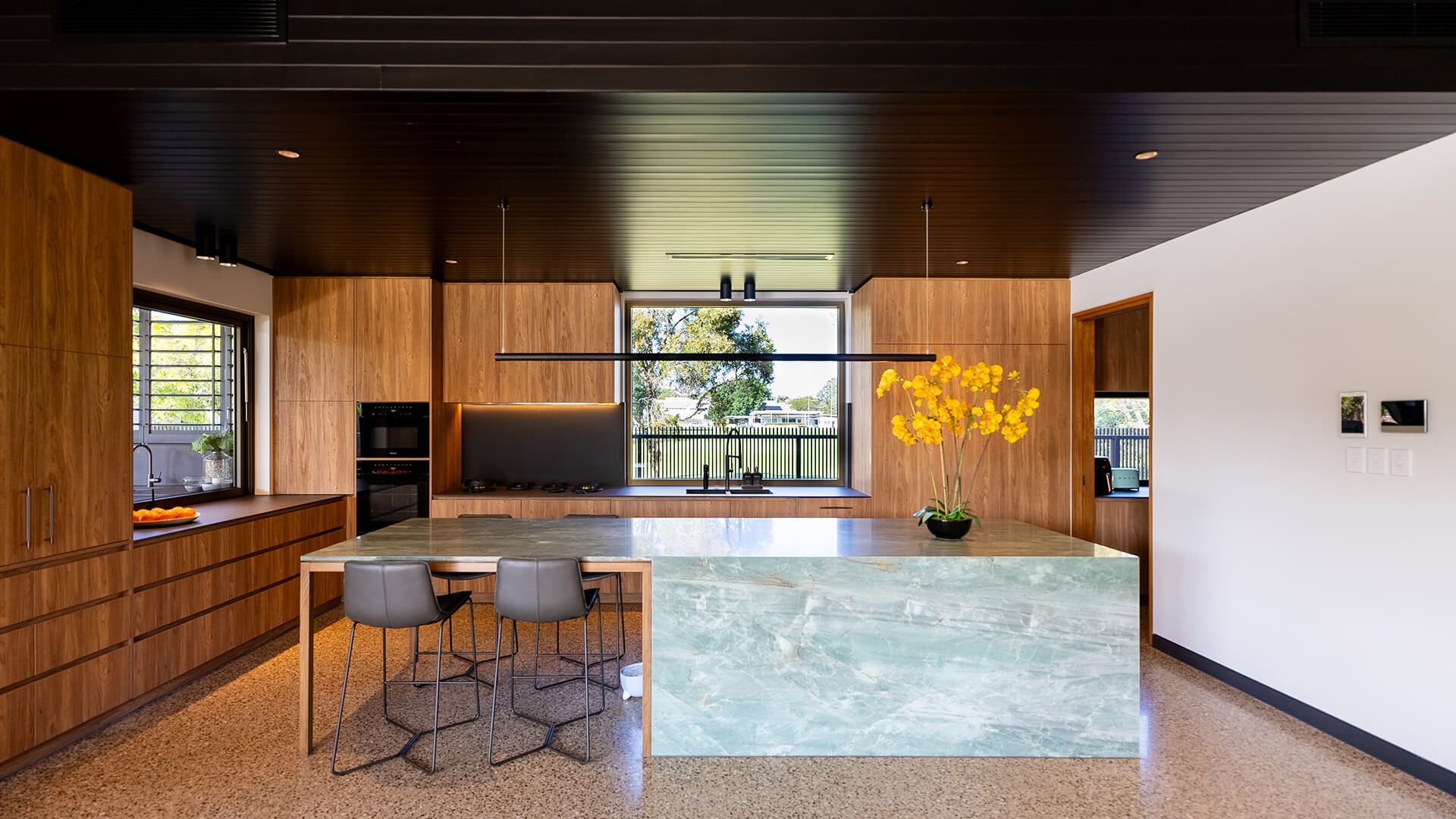 Modern kitchen with wood paneling, pale green island, stools, and a vase of yellow flowers by large windows