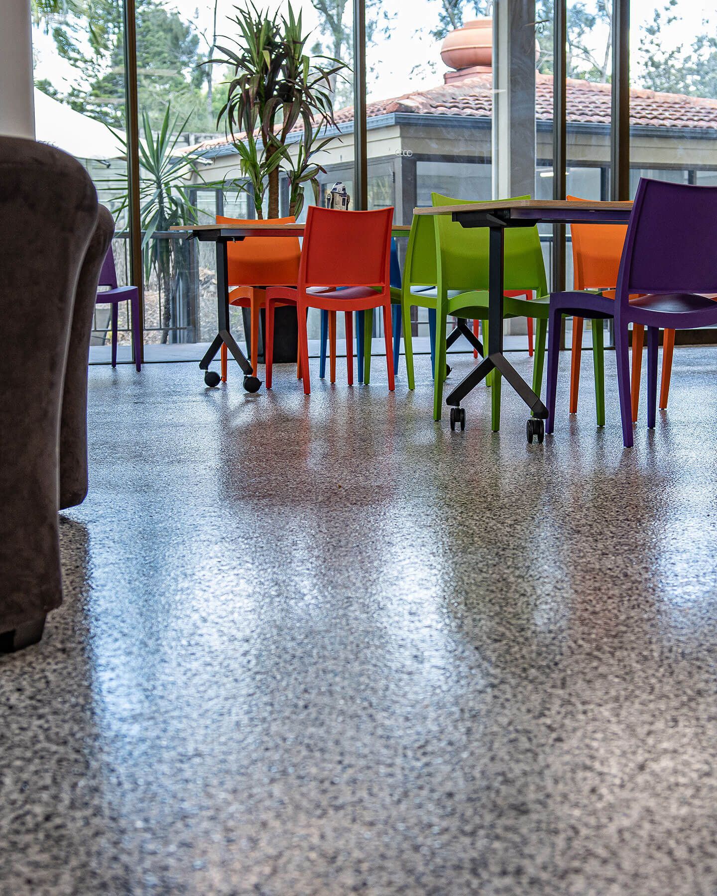 Colorful chairs and tables in a cafe by a large window, reflecting on a polished floor.