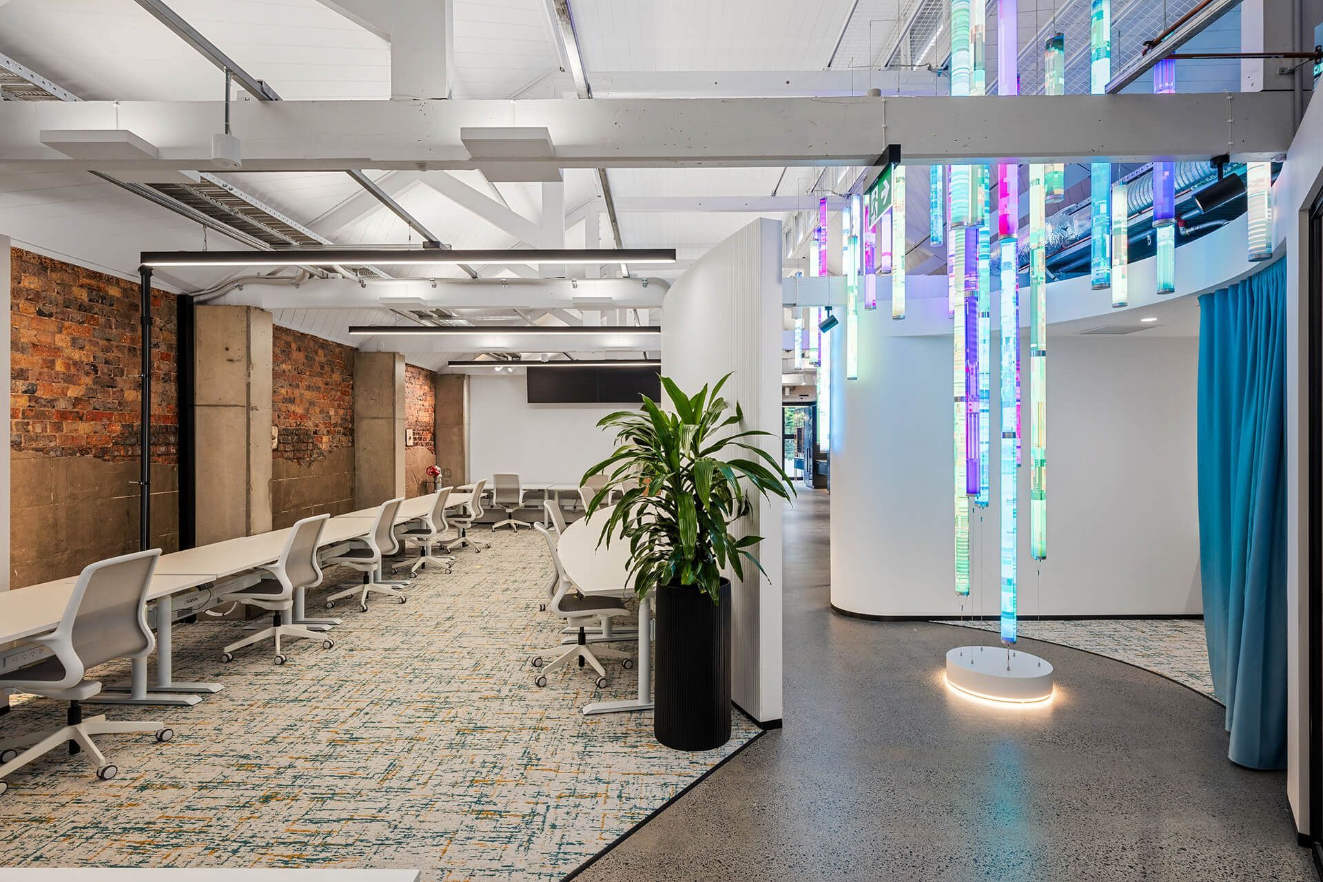 Modern office lobby with white chairs, brick wall, potted plant, and hanging colorful light sculptures.