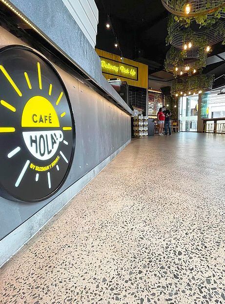 Cafe hallway with a black-and-yellow “Hola” sign, terrazzo floor, and hanging plants overhead