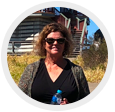 Smiling woman in sunglasses standing outdoors by stairs, holding a blue water bottle.