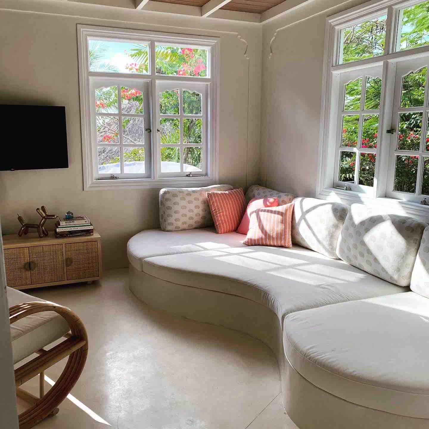 Cozy white living room with curved white sofa, windows, and colorful throw pillows. Sunlight streams in.