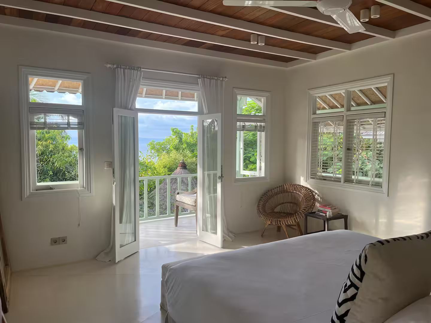 Bright bedroom with balcony overlooking greenery and the ocean. White walls, bed, and trim. Brown ceiling and wicker chair.
