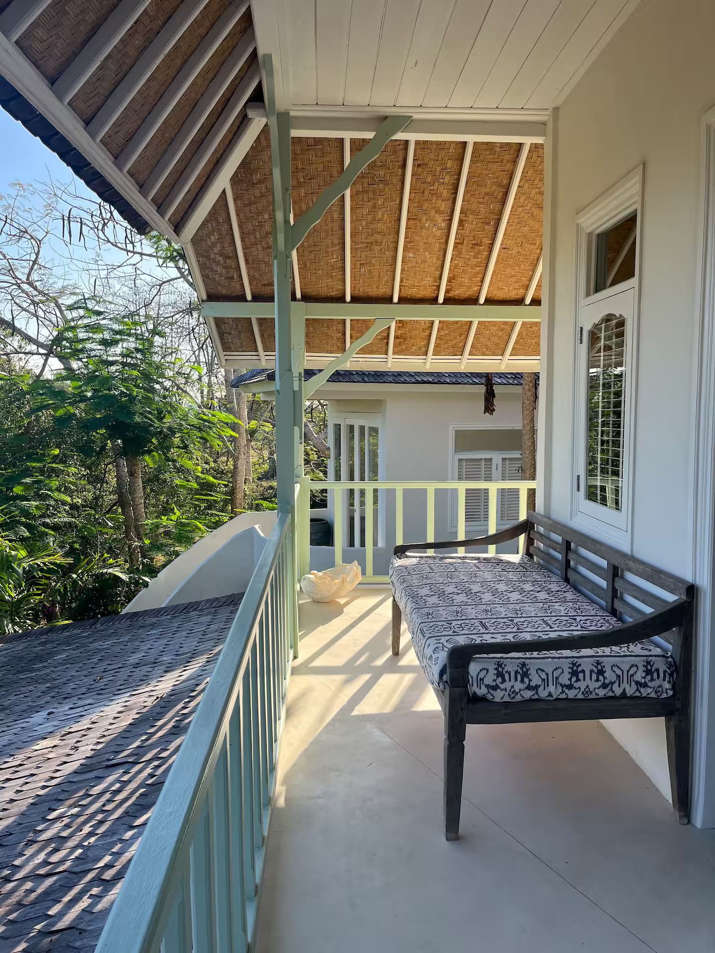 A covered porch with a wooden bench. The porch has a pale green railing and trim. Sunlight streams across the concrete floor.