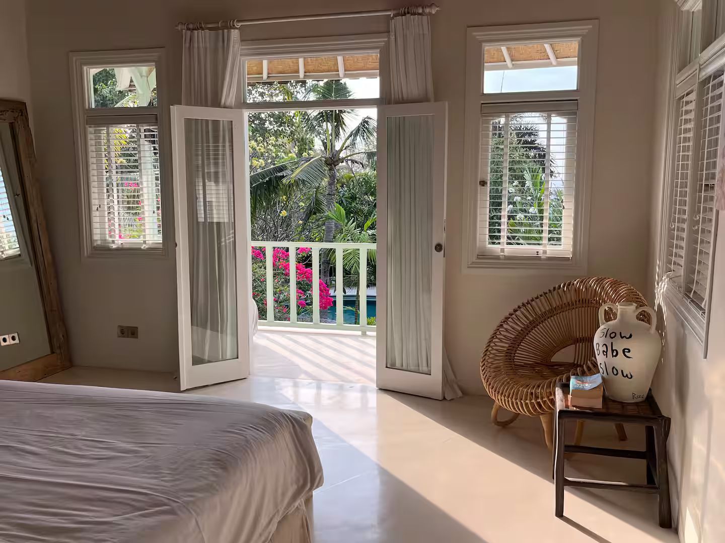 Bedroom with balcony doors open to a view of greenery and pink flowers. Includes a bed, wicker chair, and a mirror.
