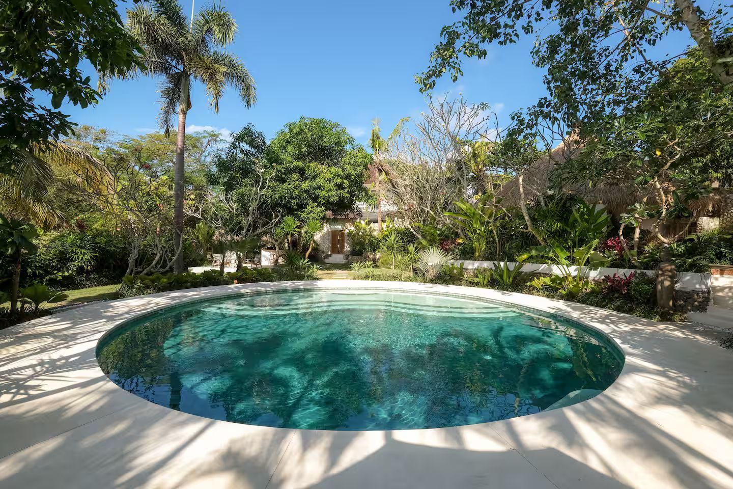Round pool surrounded by white concrete, lush greenery, and blue sky.