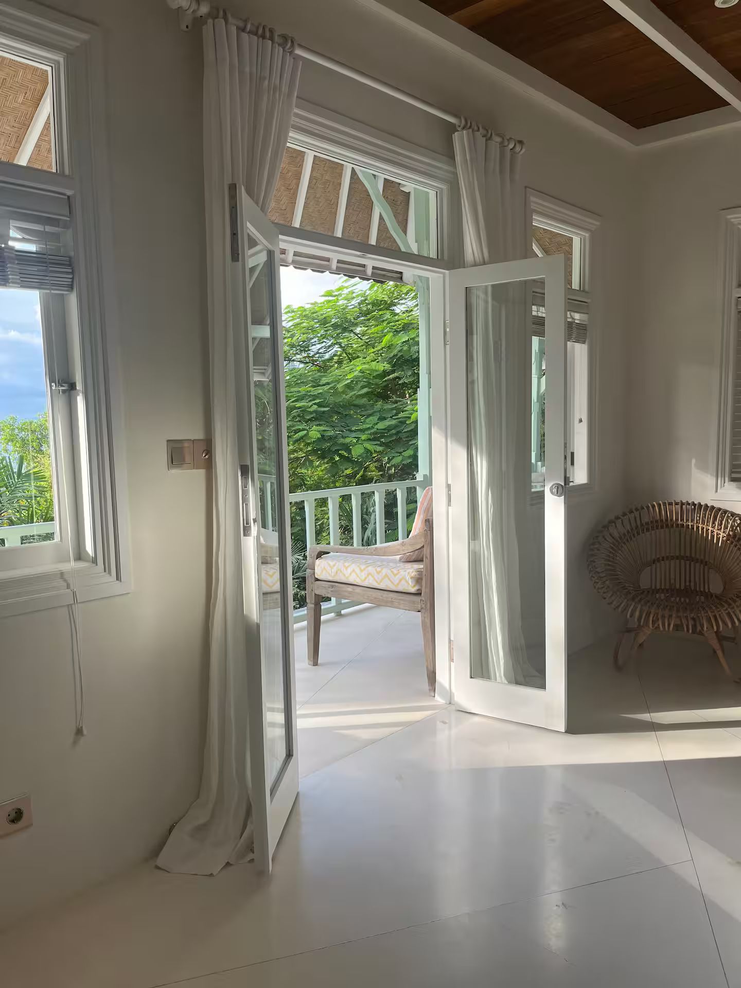 Bright room with open white French doors leading to a balcony with a chair. Sunlight streams in, illuminating the interior.