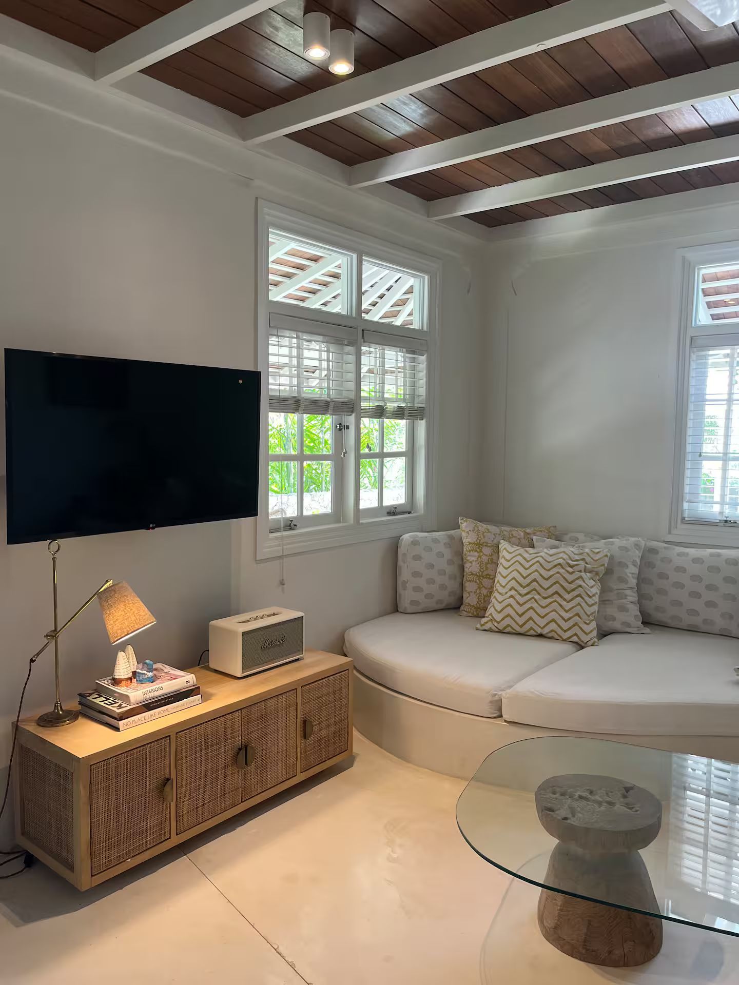 A bright living room with a curved sofa, wooden cabinet, TV, and a coffee table, next to two windows with natural light.