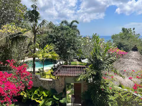 A lush, tropical garden with a pool, ocean view, and a stone archway. Bright pink flowers, green foliage, and a blue sky.