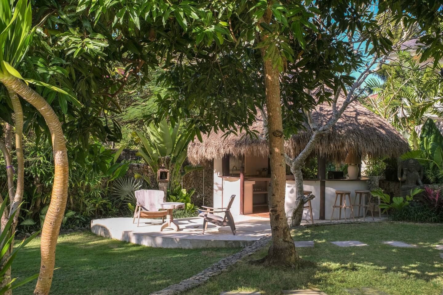 Lush garden setting with a thatched-roof outdoor bar, lawn, and seating area. Sunlight filters through the green foliage.