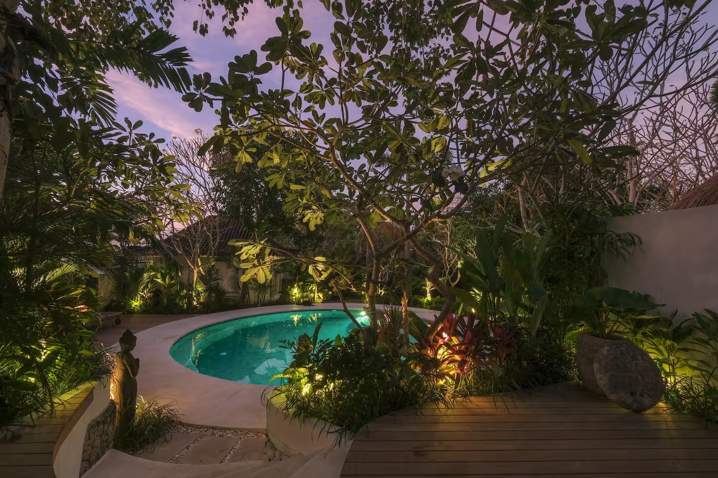 Small, illuminated pool surrounded by lush greenery and a wooden deck; twilight sky in the background.